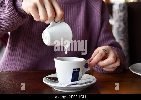 Young woman hands hold white ceramic milk jug with dripping drop into coffee cup. Pouring milk into crockery mug of warm drink with logo of Lavazza br Stock Photo