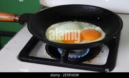 Huge chicken egg with two yolks frying in old cast pan on flaming gas blue fire of kitchen stove. Tasty homemade organic food closeup Stock Photo