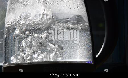 Defocused bubbles in hot water boiling inside glass teapot in bright sunlight. Closeup of boiling water in old transparent electric kettle on kitchen. Stock Photo