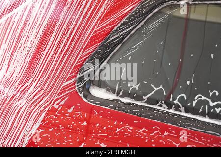 Red car washed in self serve carwash, detail on white soap foam flowing over side window and red body plates Stock Photo