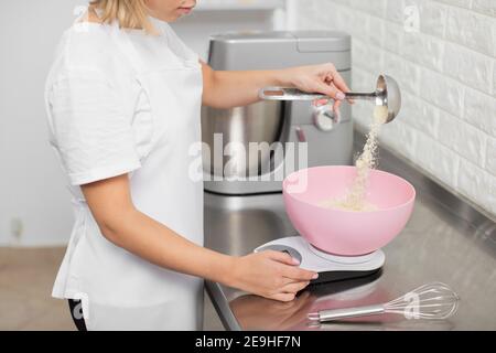 Close up cropped shot of unrecognizable woman confectioner in white apron, weighing ingredients for cookies, putting almond flour into the pink Stock Photo