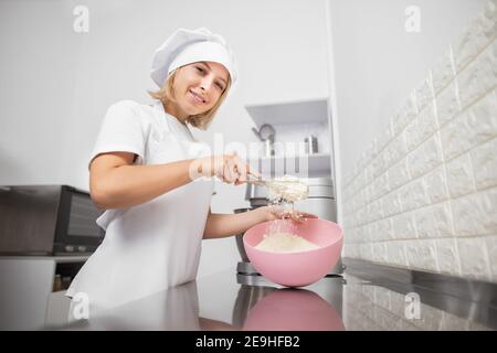 Portrait of beautiful woman pastry chef, wearing white hat and apron, mixing the flour in a pink bowl, posing to camera with smile, standing in cozy Stock Photo