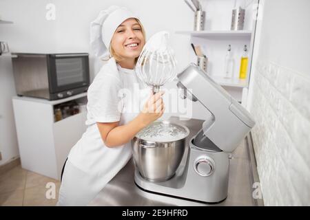 Happy pretty confectioner girl holding an electric whisk with whipping whites, while preparing a cake or desserts, using modern food processor Stock Photo