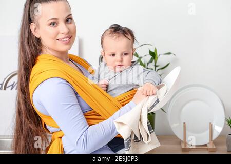 Young mother with little baby in sling washing dishes at home Stock Photo