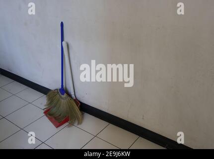 Closeup broom and dustpan standing together on tiled floor against cement wall inside a building, sweeping floor is boring work Stock Photo
