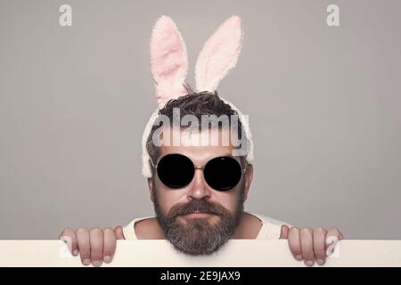 Funny man with beard in bunny ears and glasses on serious face with paper on grey background Stock Photo
