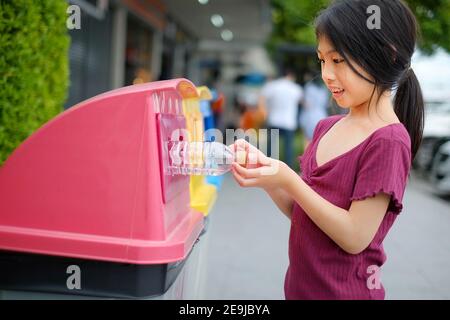 A close up picture of a cute young Asian girl throwing away an empty plastic water bottle on a recycling bin which sorted waste into 3 groups, recycla Stock Photo