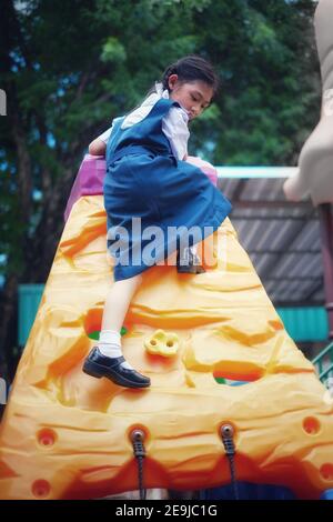 A cute young Asian girl in white and blue school uniform is playing rock climbing at a playground, exercising and having fun. Stock Photo