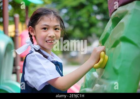 A cute young Asian girl in white and blue school uniform is playing rock climbing at a playground, exercising and having fun. Stock Photo