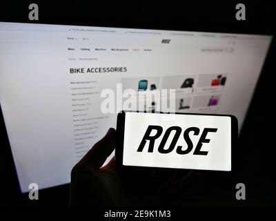 Person holding cellphone with logo of German bicycle manufacturer Rose Bikes GmbH on display in front of company website. Focus on smartphone screen. Stock Photo