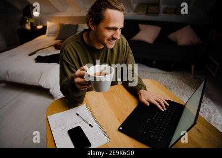 Caucasian male student enjoying hot coffee while logging onto online classes typing on laptop sitting at desk in home office Stock Photo
