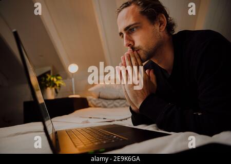 Caucasian male lying on bed concentrating while listening to online video call with colleagues Stock Photo