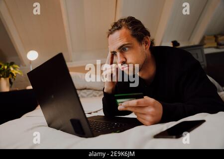 Stressed caucasian male student holding credit card making online payment using laptop lying on bed Stock Photo