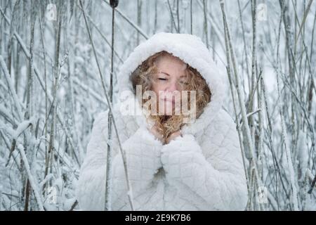 red-haired mature woman with white hooded coat stands smiling contentedly in the winter cold in the snowy reeds Stock Photo