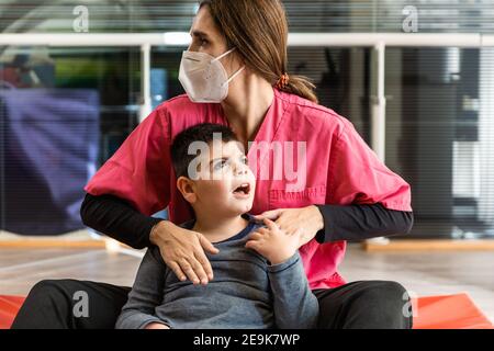disabled child and physiotherapist on a red gymnastic mat doing exercises. pandemic mask protection Stock Photo