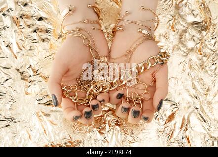 Female hands with trendy dark nail design with gold bracelets on golden background. Luxury concept. Festive backdrop for your design. Top view. Stock Photo