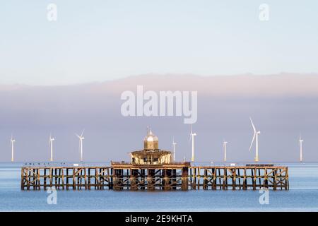 The abandoned Victorian pier head off the coast of Herne Bay in Kent, England. The pier head is over a mile out and surrounded by a very calm sea on a winters day with clear blue skies and a layer of cloud on the horizon. Directly behind the pier are the white wind turbines of the London array windfarm Stock Photo