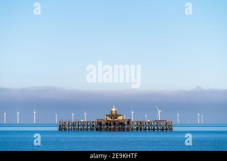 The abandoned Victorian pier head off the coast of Herne Bay in Kent, England. The pier head is over a mile out and surrounded by a very calm sea on a winters day with clear blue skies and a layer of cloud on the horizon. Directly behind the pier are the white wind turbines of the London array windfarm. Stock Photo