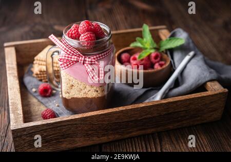 No-bake layered dessert with dark chocolate mousse, crushed cookies and raspberry cream cheese in glass jar on rustic wooden background Stock Photo