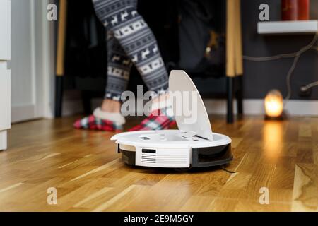 Caucasian young man in christmas pajamas doing vacuum cleaner robot maintenance. Male in winter patterned sleepwear and red socks slippers starts Stock Photo