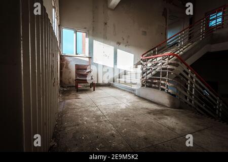 Abandoned building creepy dark moody staircase in dilapidated run down old deserted hospital school ruin with a single empty chair indoors on stairs Stock Photo