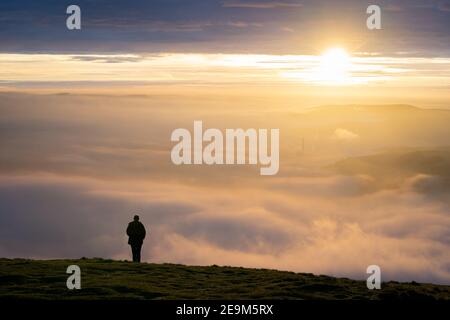 Silhouette of one lone man stood on edge of mountain peak top above clouds as fog mist and cloud inversion gathers watching the sun rise a beautiful Stock Photo