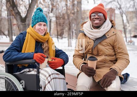 Portrait of young happy couple smiling at camera while sitting in winter park and drinking hot coffee Stock Photo