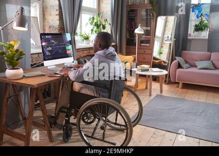 Rear view of African man sitting on wheelchair at the table in front of computer monitor and working with software at home Stock Photo