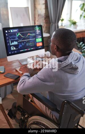 Rear view of African man sitting on wheelchair typing on computer keyboard and working with computer program at home Stock Photo
