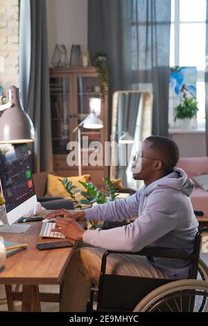 African graphic designer sitting on wheelchair in front of computer monitor working online at home Stock Photo