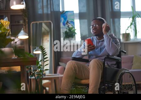 African disabled man in wireless headphones using mobile phone to listen to music while sitting on wheelchair in the room Stock Photo