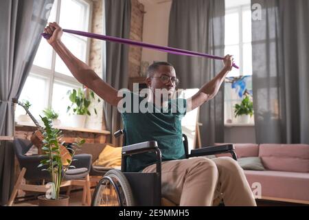 African disabled man sitting in wheelchair raising his arms and doing stretching exercises during sports training at home Stock Photo
