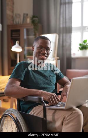 Portrait of African disabled man sitting in wheelchair with laptop on his knees and smiling at camera working at home Stock Photo