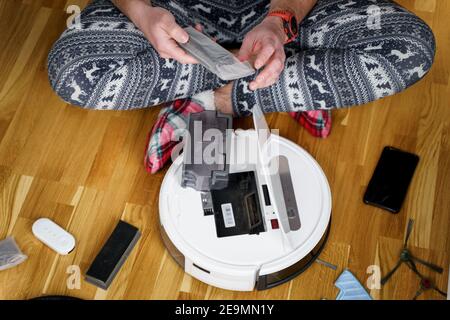 Caucasian young man in christmas pajamas doing vacuum cleaner robot maintenance. Male in winter patterned sleepwear and red socks slippers starts Stock Photo
