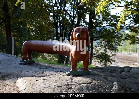 Long dachshund shaped brown bench located in a dog park in Helsinki, Finland. Photographed Sep, 2019. The dog shaped bench is on a rock front of trees Stock Photo
