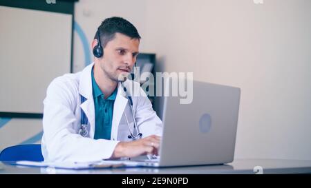 Friendly Male Doctor in White Medical Coat With Headphones Making Conference Call on Laptop.Remote Consulting Patient Online From Healthcare Hospital Stock Photo
