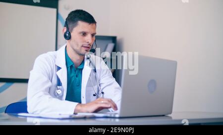 Friendly Male Doctor in White Medical Coat With Headphones Making Conference Call on Laptop.Remote Consulting Patient Online From Healthcare Hospital Stock Photo