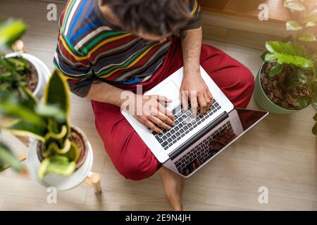 Young man working at home with laptop, sitting on the floor surrounded by houseplants Stock Photo