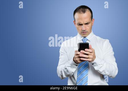 Mature Brazilian man, in formal clothes, holding his smartphone with a serious face. Blue background. Stock Photo