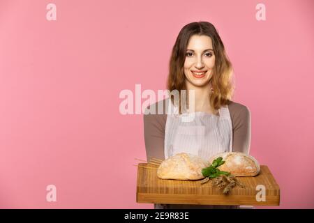 A young girl stands on a light pink background and in her hand she holds a board with two loaves of bread decorated with herbs and ears of cereals... Stock Photo