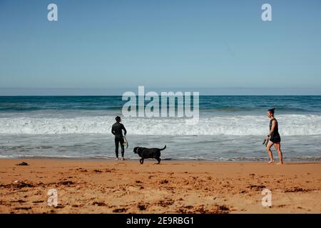A surfer looks out the waves as a a lady walks her dog on the beach of the Southern Ocean at Moyji or Point Ritchie in Warrnambool, Southwest Victoria Stock Photo