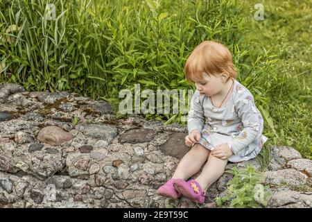 Little girl with red hair sits with a camomile in her hand in the garden among the greenery. Selective focus. Stock Photo