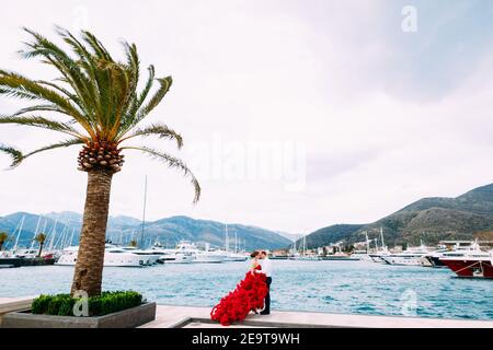 A man and a woman in a red gown are hugging on a boat pier Stock Photo
