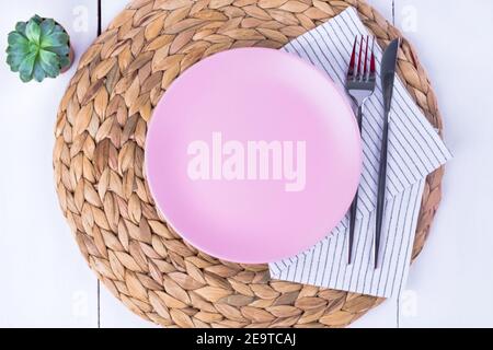 Close-up top view of a serving of empty pink plates, a knife and a fork on a straw eco-friendly napkin. Selective focus. Mockup, minimalism. Stock Photo