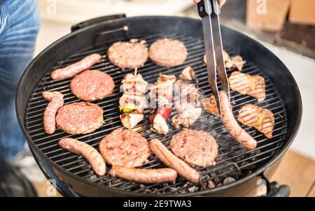 Close up of hand grilling meat at barbecue session - Skewers and burger sausages cooked on ember at bbq garden party - Pic nic concept Stock Photo