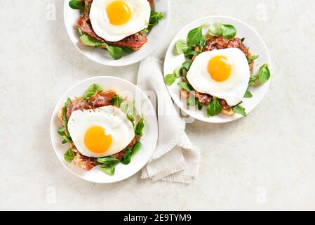 Open sandwiches with fried eggs, bacon and vegetable leaves on plates over light stone background. Top view, flat lay Stock Photo