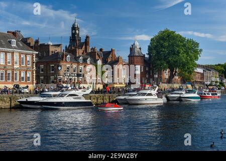 People relax in sun outside busy riverside pub & leisure boats moored on water at River Ouse quayside - King's Staith, York, North Yorkshire, England. Stock Photo