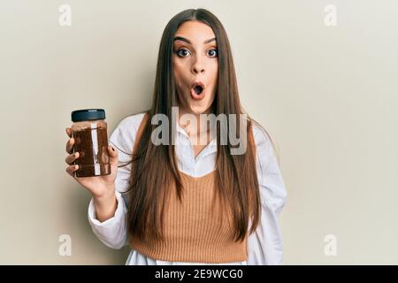 Beautiful brunette young woman holding soluble coffee scared and amazed with open mouth for surprise, disbelief face Stock Photo