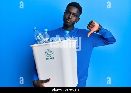 Young african american man holding recycling wastebasket with plastic bottles with angry face, negative sign showing dislike with thumbs down, rejecti Stock Photo