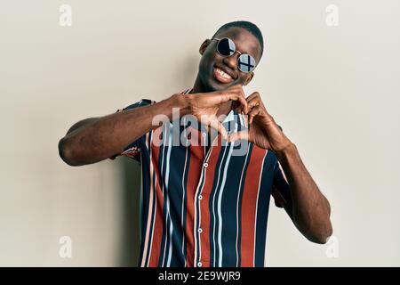 Young african american man wearing casual clothes and sunglasses smiling in love doing heart symbol shape with hands. romantic concept. Stock Photo